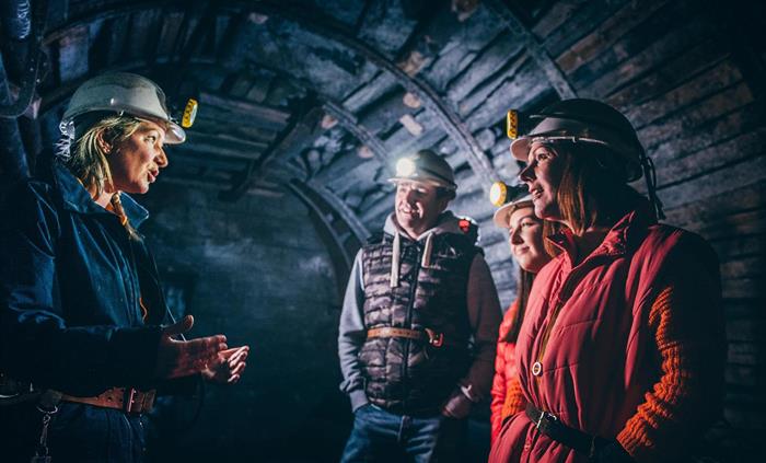 A guide is dressed in dark blue overalls and a white hard hat. She is talking to three visitors, a man, woman and teenage girl. They are also wearing